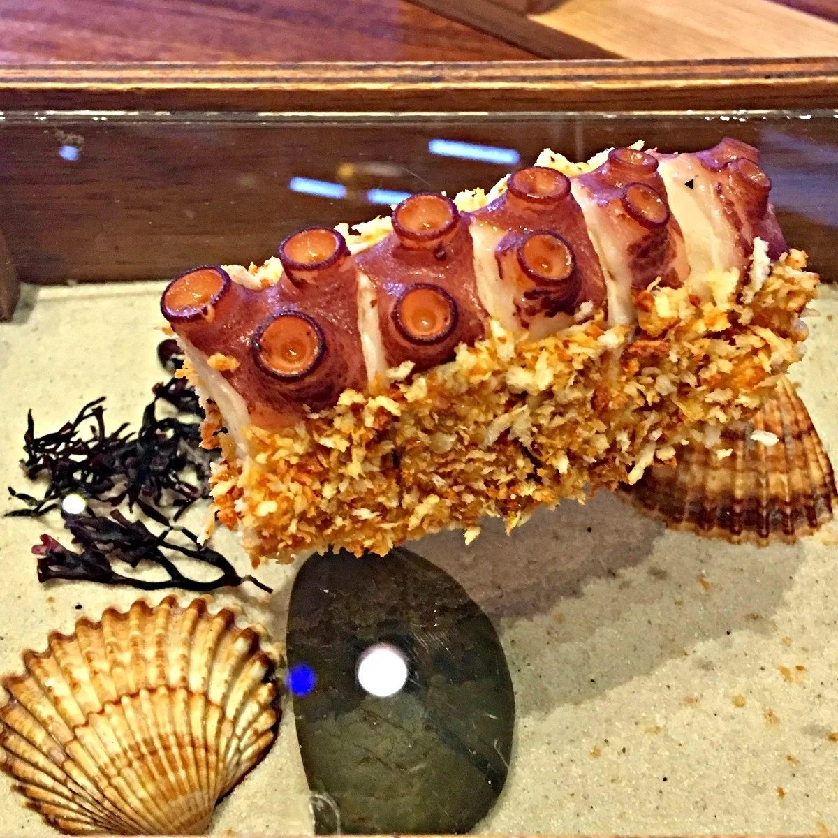 Tentáculo de pulpo rebozado en panko presentado sobre arena con conchas y alga seca en Tickets